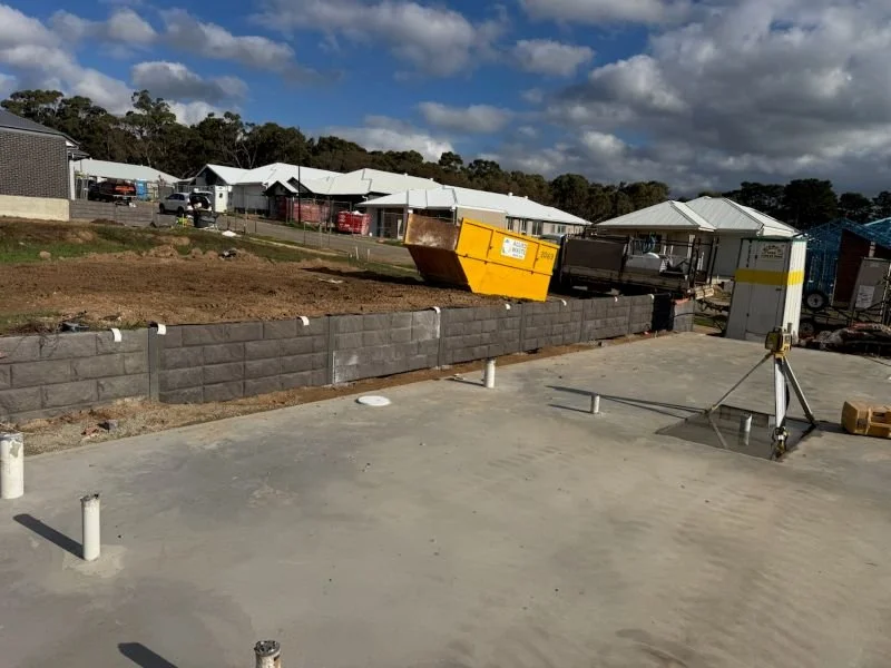 Construction site with a cinder block wall in progress, a concrete slab, construction equipment, and residential houses in the background under a partly cloudy sky.
