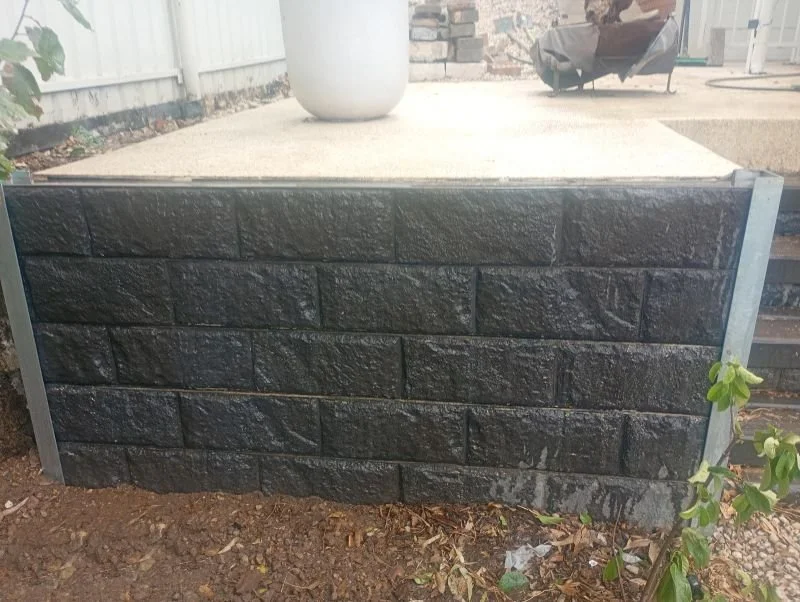 Black landscaped retaining wall with concrete surface on top and a potted plant on the patio.
