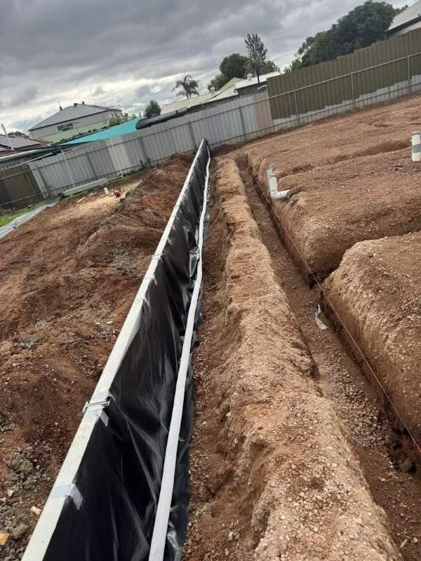 Construction site with trenches and foundation pipes, enclosed by a metal fence, overcast sky.