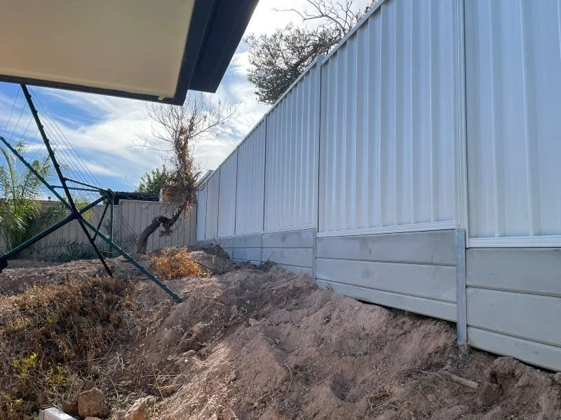 A backyard with a white metal fence, leaning on uneven dirt terrain with rocks, and an outdoor twisted metal chair or swing in the corner. The sky is partly cloudy, and there are some trees in the background.