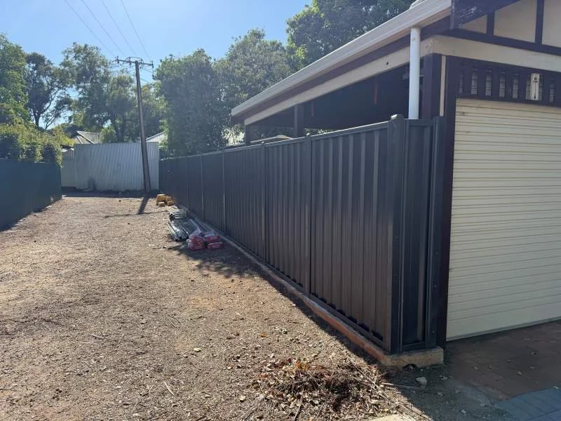 A black metal fence installed along a property with a garage on the right, a dirt ground, some construction materials, and trees in the background.