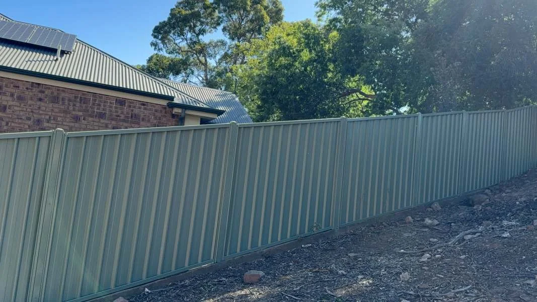 Image of a corrugated metal fence on a sloped ground in front of a brick house with a metal roof and solar panels on top, surrounded by green trees.