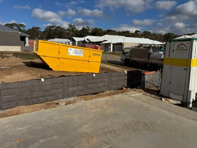 Construction site with an orange temporary waste container labeled 'Allied Waste,' a partially built black brick wall, a small white utility door with a yellow stripe, and a construction truck parked in the background. Residential houses and a blue s