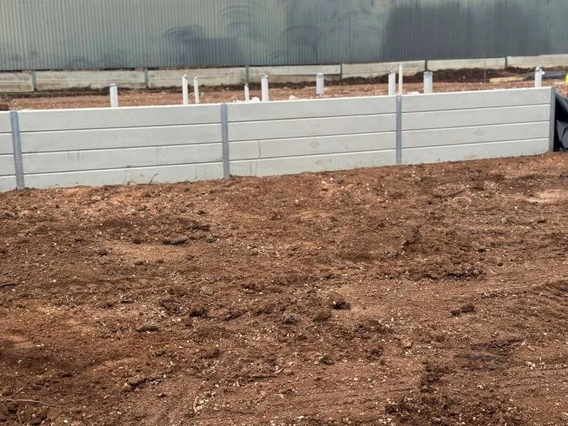 A dirt ground with a partially built raised garden bed made of white panels, supported by vertical metal stakes, with a background of a concrete or metal wall.