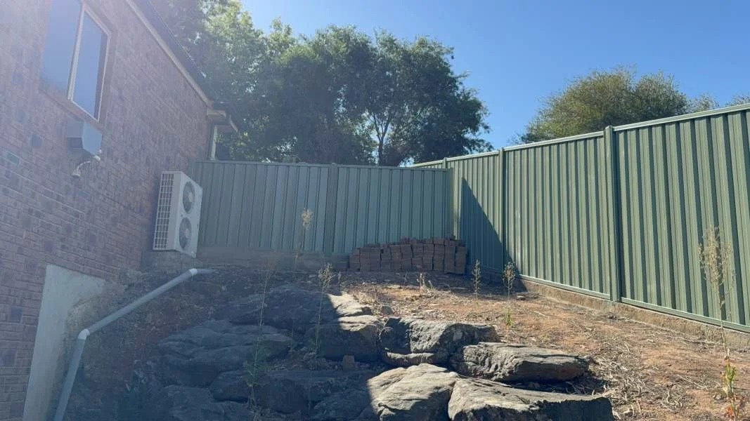Backyard with rocky slope, green metal fence, and brick wall with HVAC unit