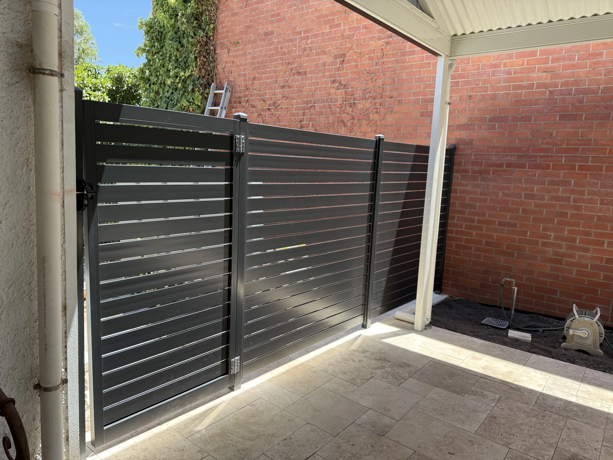 View of a small backyard patio with a black metal fence, a brick wall, a ladder, and some construction tools.