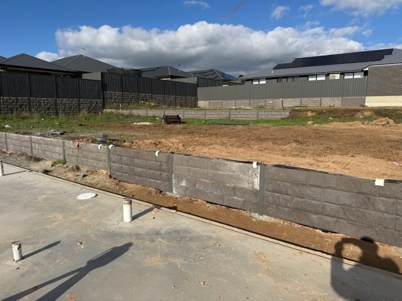 Construction site with a partially built retaining wall, dirt ground, and surrounding fences, in a suburban neighborhood with houses and cloudy sky.