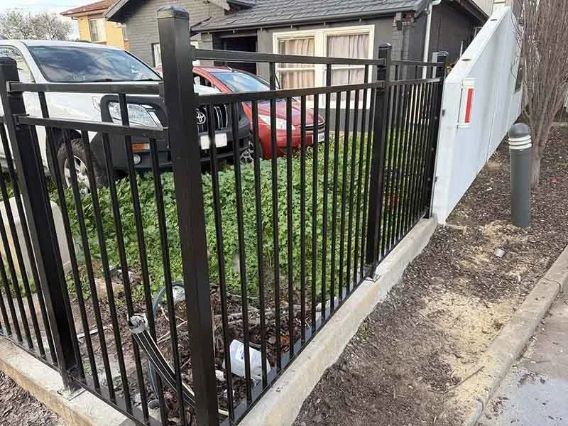 A black metal fence with vertical bars surrounds a small landscaped area with green shrubbery. Behind the fence, there are parked cars, including a white SUV and a red sedan, and a gray building with a window and a sloped roof. On the right side, the