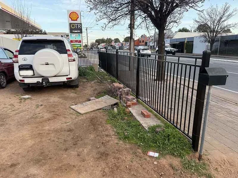 A white SUV parked on dirt next to a black metal fence along a sidewalk, with a Shell gas station sign and restaurant in the background.