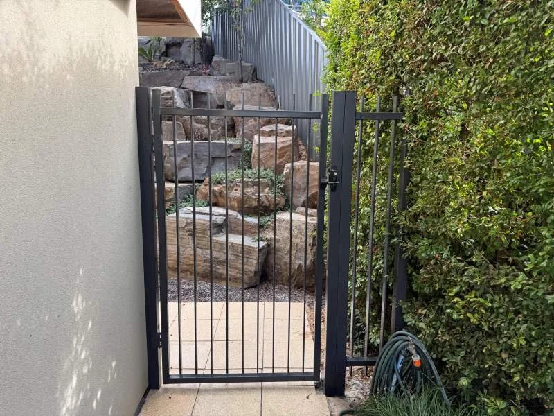 Black metal gate with loose latch, leading to a rock garden with large rocks and a small slope, bordered by a green hedge.