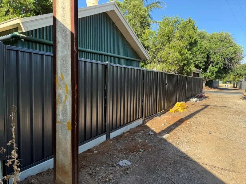A black metal fence running along a dirt pathway in a residential area with a house and trees in the background.
