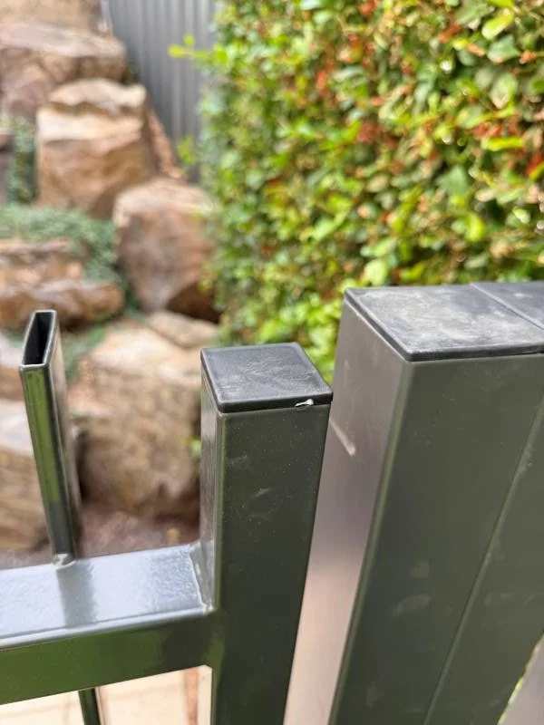Close-up of a metal gate with a vertical bar and a rectangular post, with a stone rock formation and green bushes in the background.