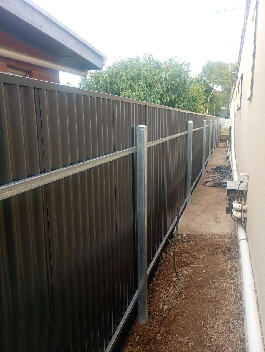 Side view of a newly installed metal fence running along the side of a house with a small walkway and plumbing pipes visible on the right.
