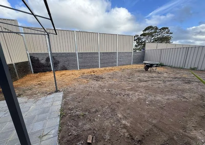 Empty backyard with dirt ground, a small paved patio area, a wheelbarrow, and a goodneighbour fence on retaining wall, under a partly cloudy sky.