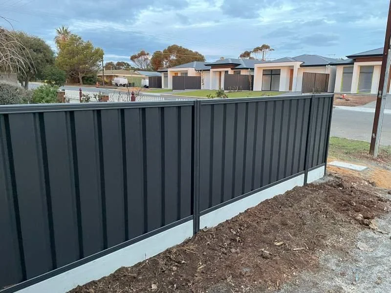 A newly installed black metal fence runs along a residential property with modern houses in the background, under a partly cloudy sky.