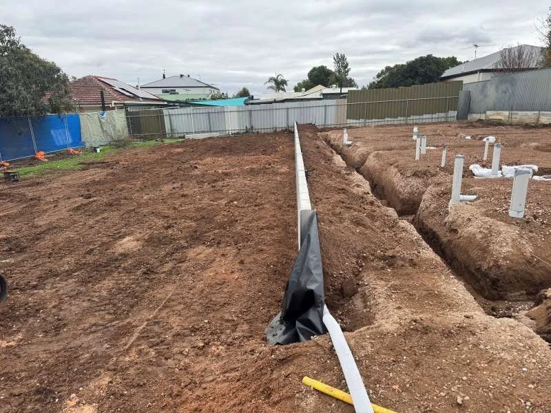 Construction site with trenches and plumbing pipes for foundation work, dirt ground, houses and trees in background.