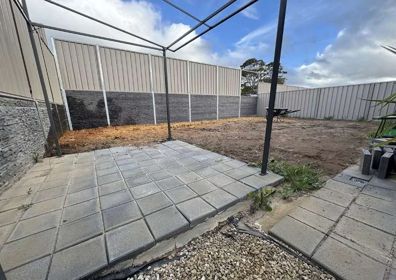 Backyard with partially paved patio, dirt area, and a goodneighbour on retaining wall fenced enclosure. Metal poles indicate a frame for a structure like a shed or canopy, with some construction materials on the side.