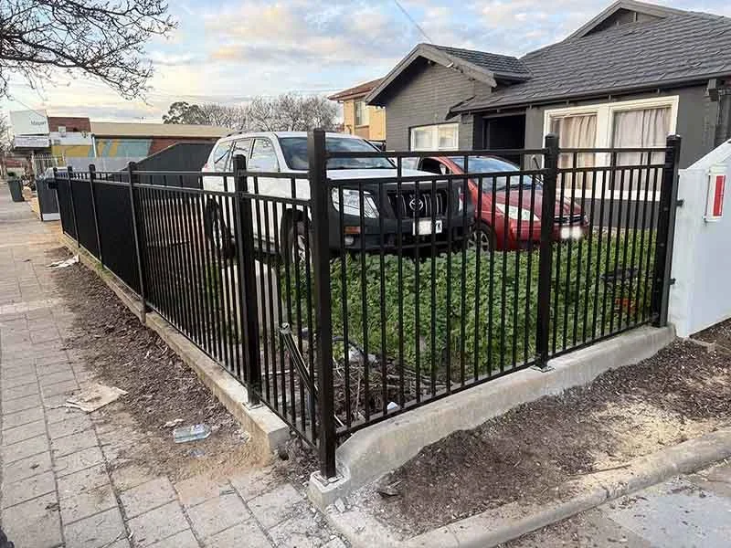 A black metal fence surrounds a small patch of grass with two parked cars, a white SUV and a red sedan, beside a house with gray siding and white curtains.