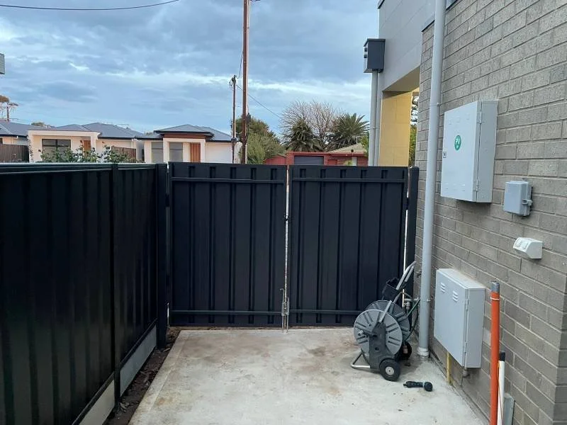Small backyard area with a concrete floor, enclosed by black metal fencing, with a black gate, an orange pipe, and utility boxes on the house wall. A hose reel and an outdoor power tool are leaning against the house wall, and the sky is cloudy.