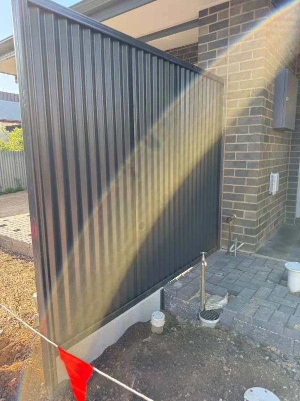 Outside backyard scene showing a dark gray metal fence, a brick house wall, and some pipes and construction materials on the ground with a construction flag in the foreground. A sunbeam shines across the scene.