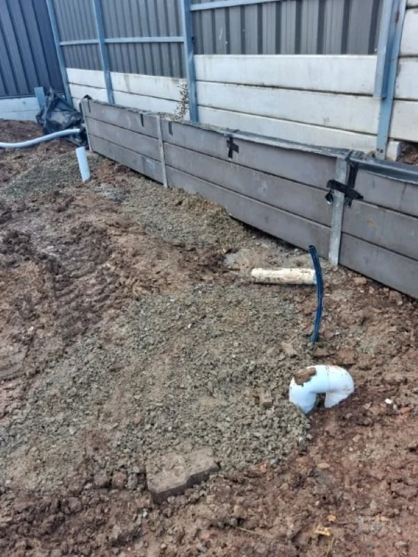 Construction site showing a trench with soil and exposed pipes, and a partially installed wooden fence along a corrugated metal fence.