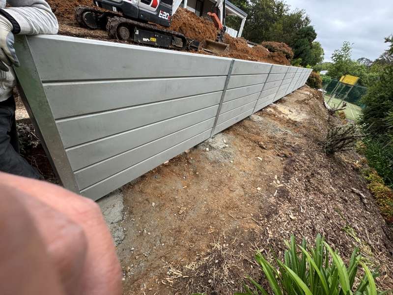 Construction site with a new gray metal retaining wall, dirt ground, and a small excavator on top of the dirt pile. There are trees and a tennis court fence in the background.
