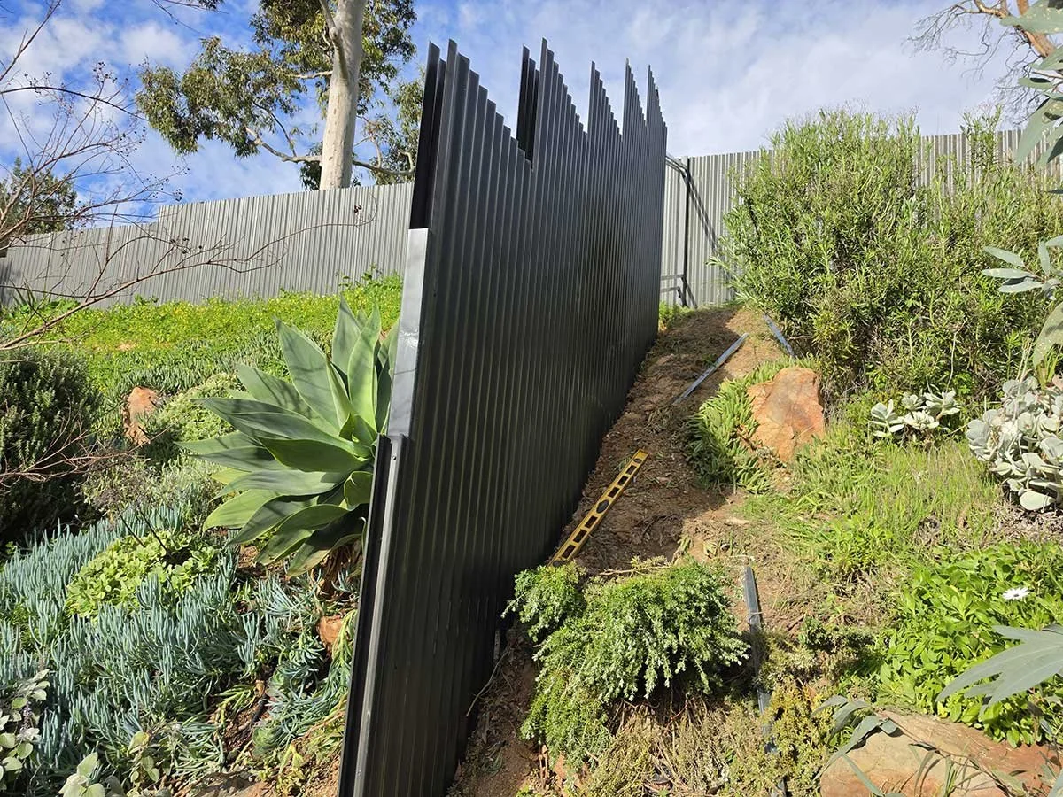 Post +rail twin fence in a very slope land grey color around green bushes