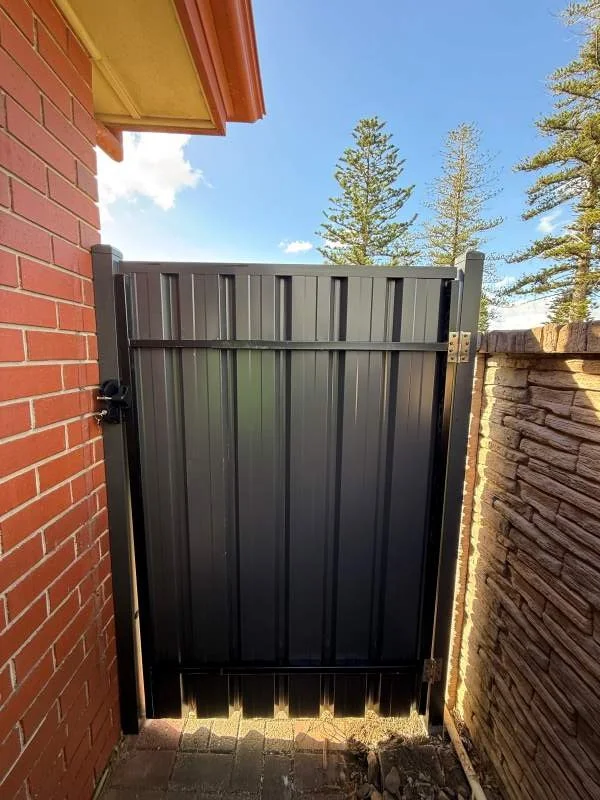 A black metal gate between a red brick wall on the left and a stone wall on the right, with a cloudy sky and trees in the background.