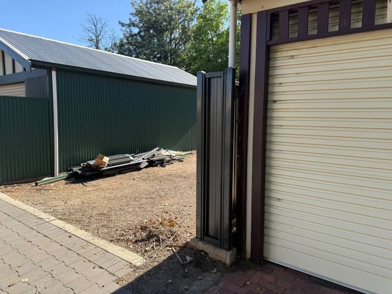 Side view of a partially open garage door with a yellow roll-up door next to a small side gate, and a yard with dirt and debris on a sunny day.