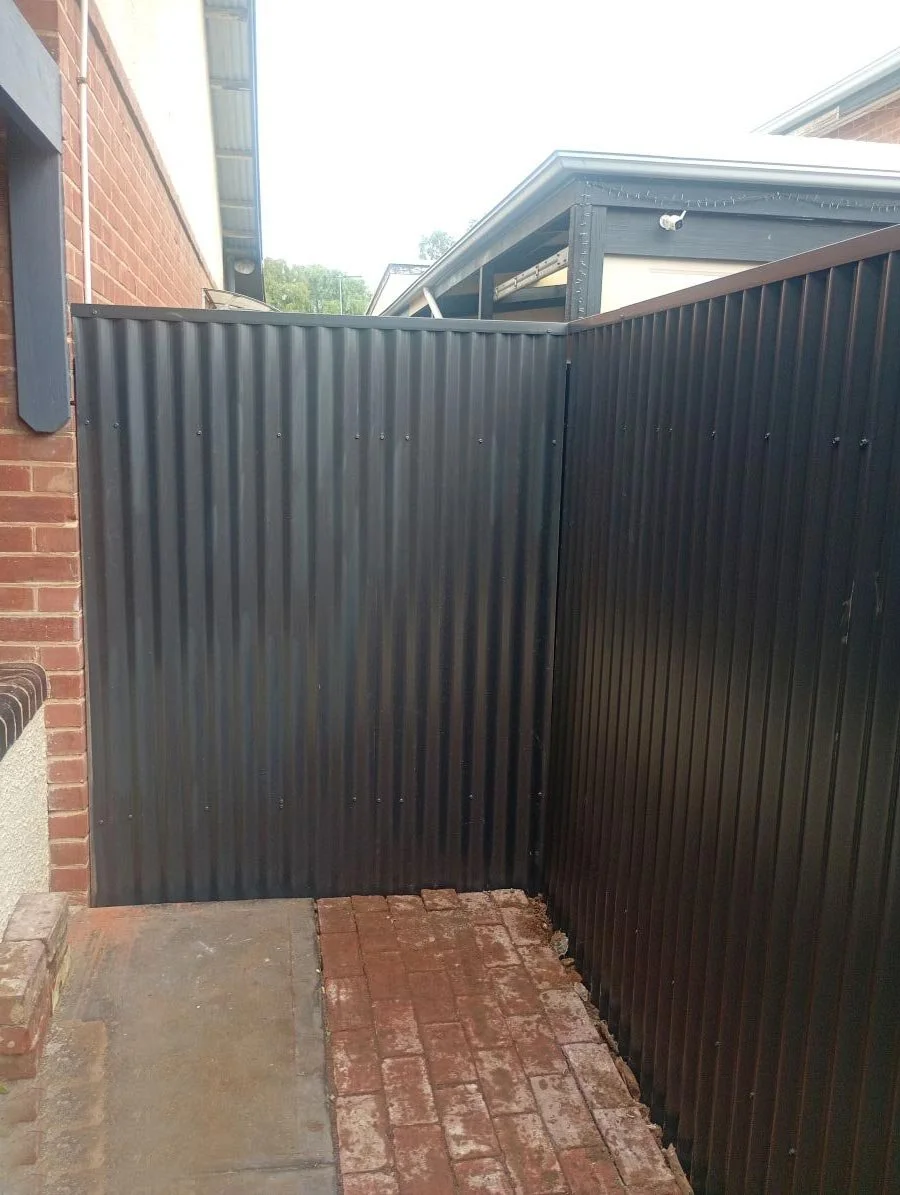A corner with black corrugated metal fencing next to a brick wall and brick paved walkway.