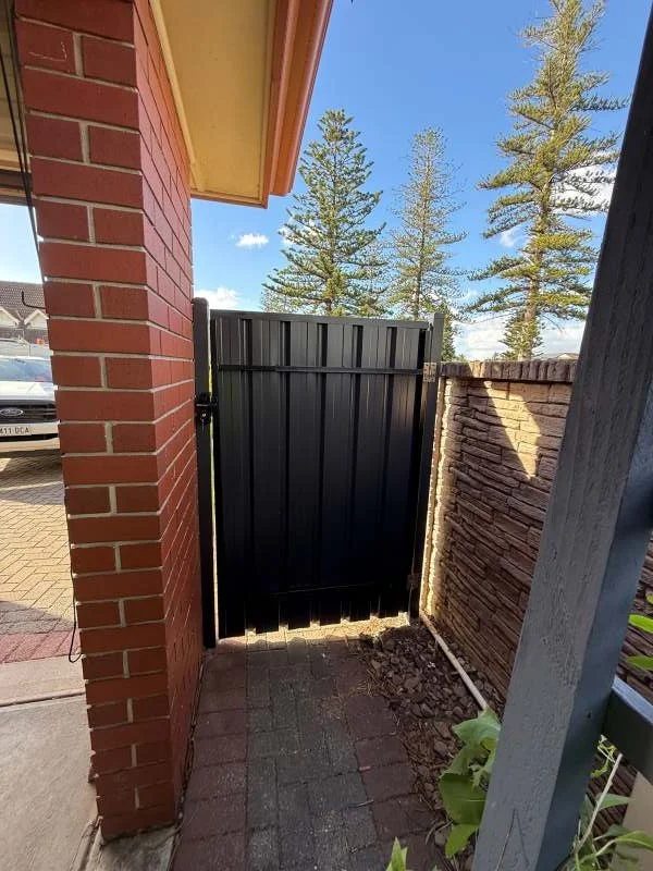 View of a small outdoor entrance area with a brick wall on the left, a black Pedestrian gate in the center, and a brick wall on the right. There are apple trees and a clear blue sky with some clouds in the background.