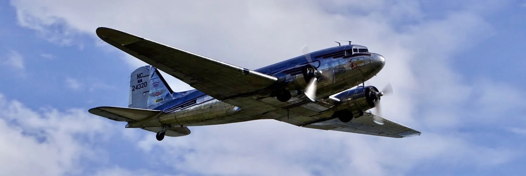 A vintage silver twin-engine propeller airplane flying in a partly cloudy sky.