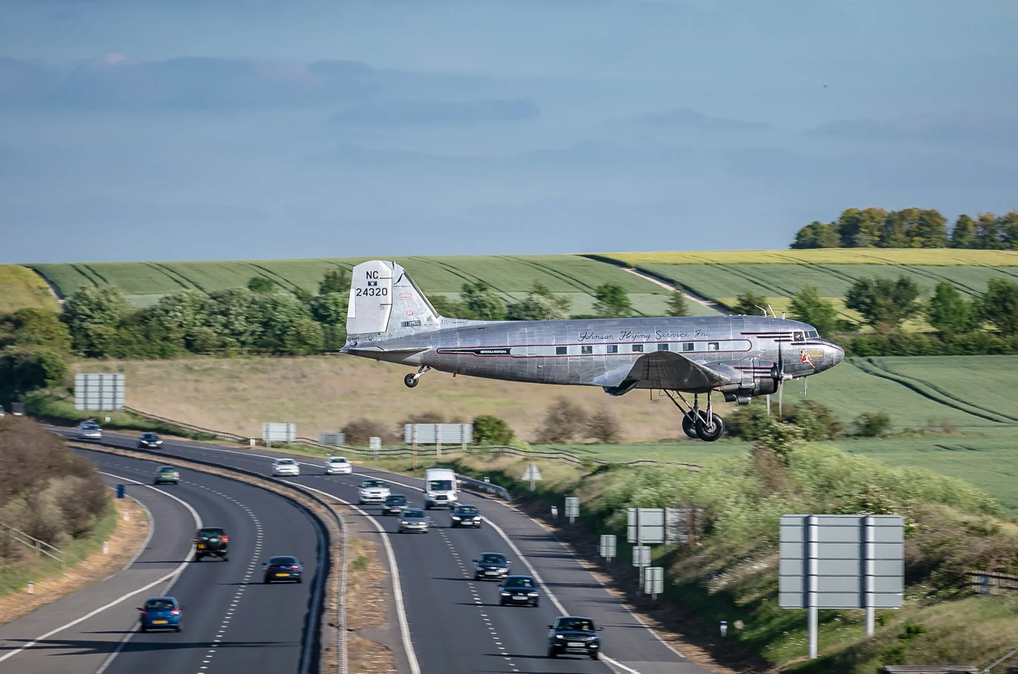 An airplane flying low over a highway with moving cars, green fields, and trees in the background under a cloudy sky.