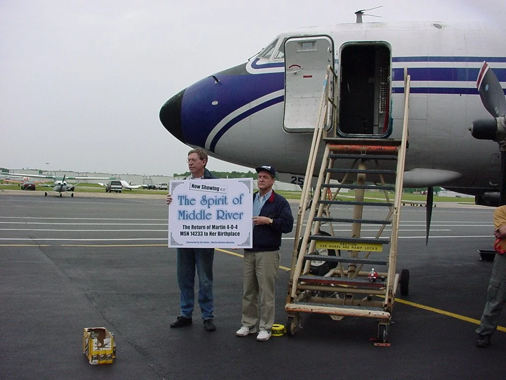Two men standing in front of an airplane on an airport tarmac, holding a sign that reads: "Now Showing The Spirit of Middle River, The Return of Martin 4-0-4, MSN 14233 to Her Birthplace."