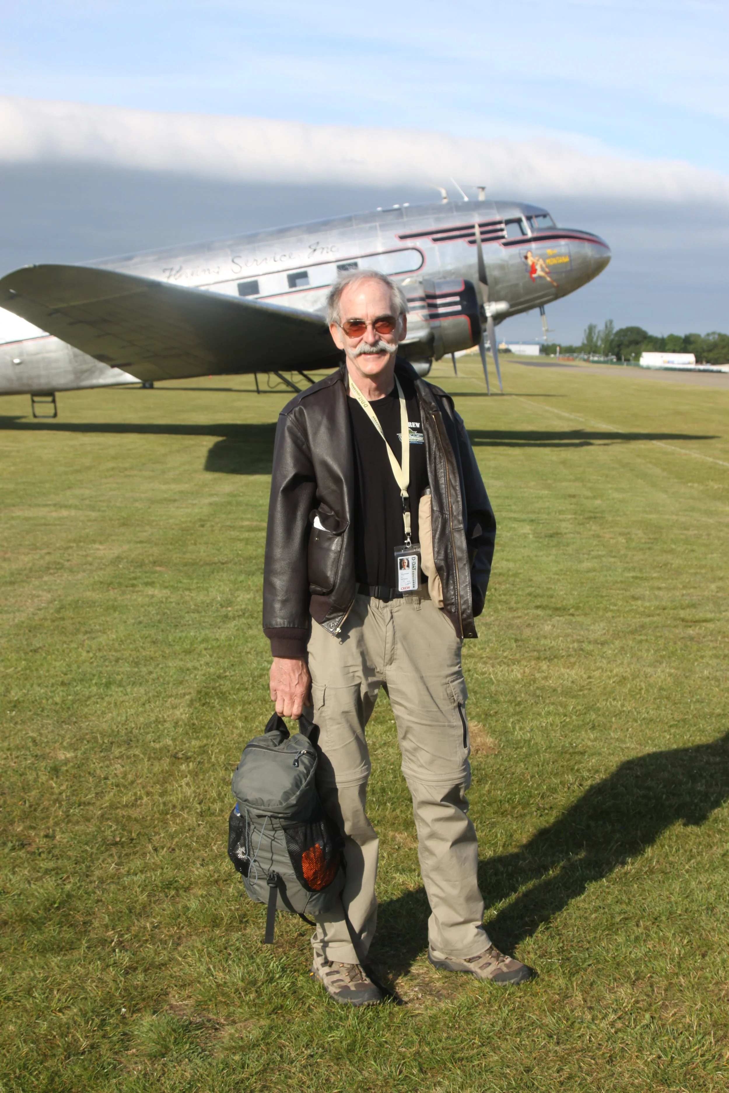 A man standing on grass next to a vintage silver airplane with a person in the background.