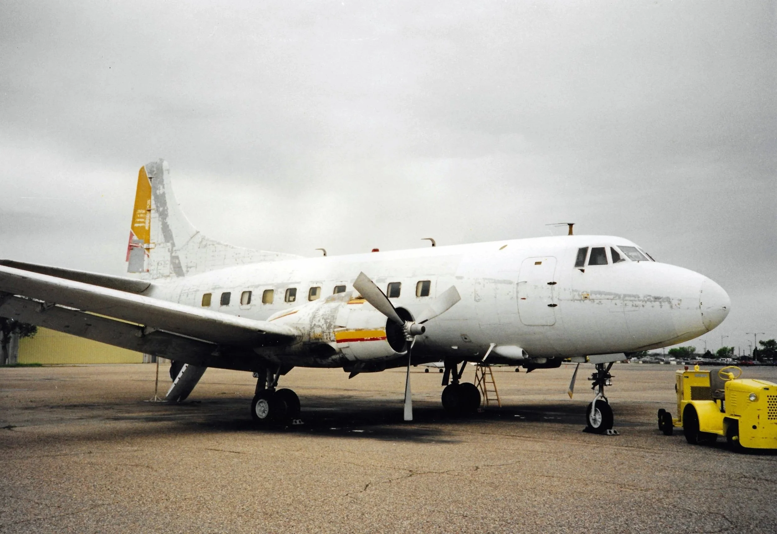 A white airplane with a yellow tail parked on a tarmac, with a cloudy sky in the background and maintenance equipment nearby.