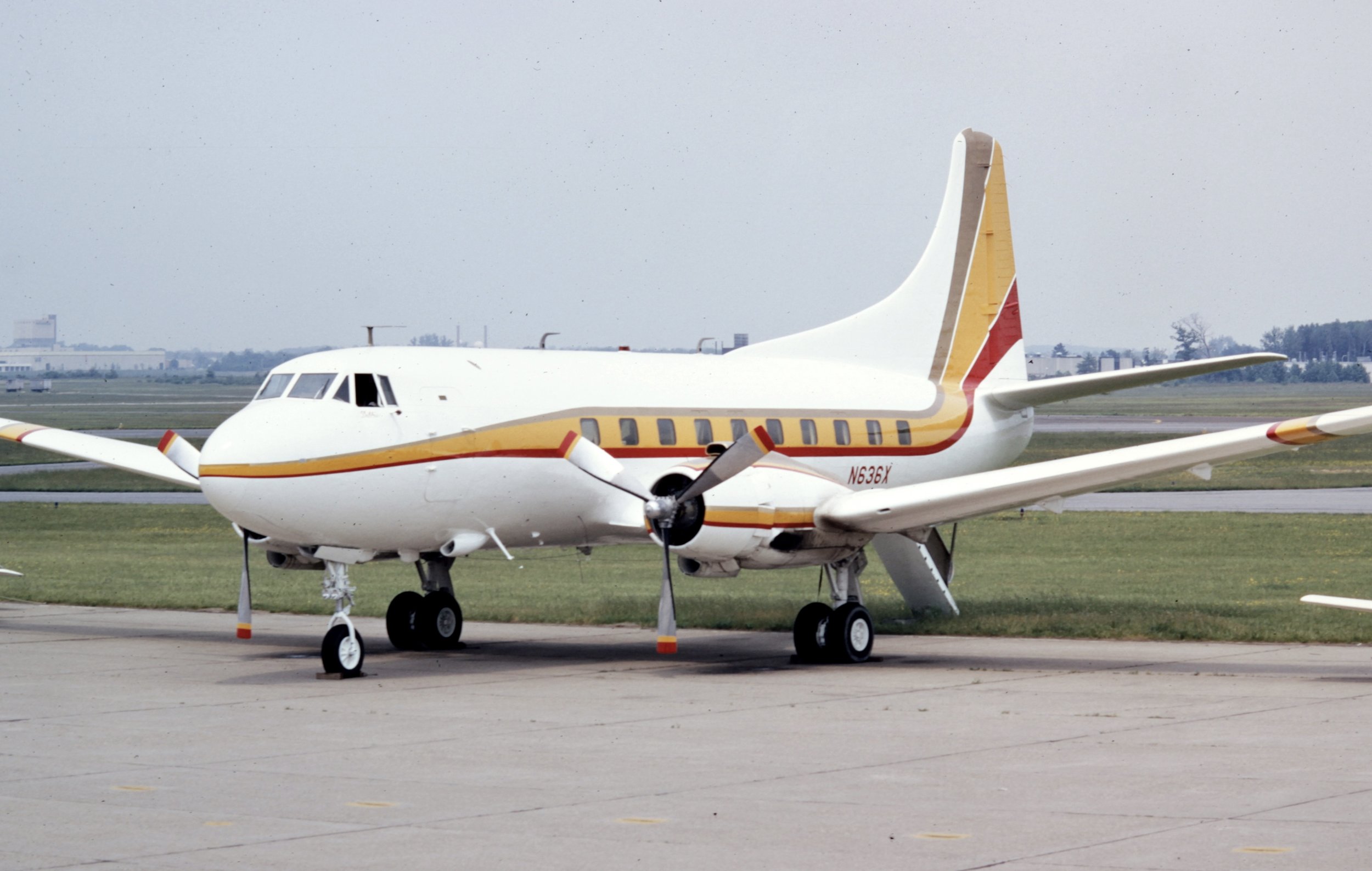 A Martin 4-0-4 airplane parked on an airport tarmac with a colorful stripe design along the side, yellow, red, and beige. The background shows a mostly overcast sky and some distant buildings.