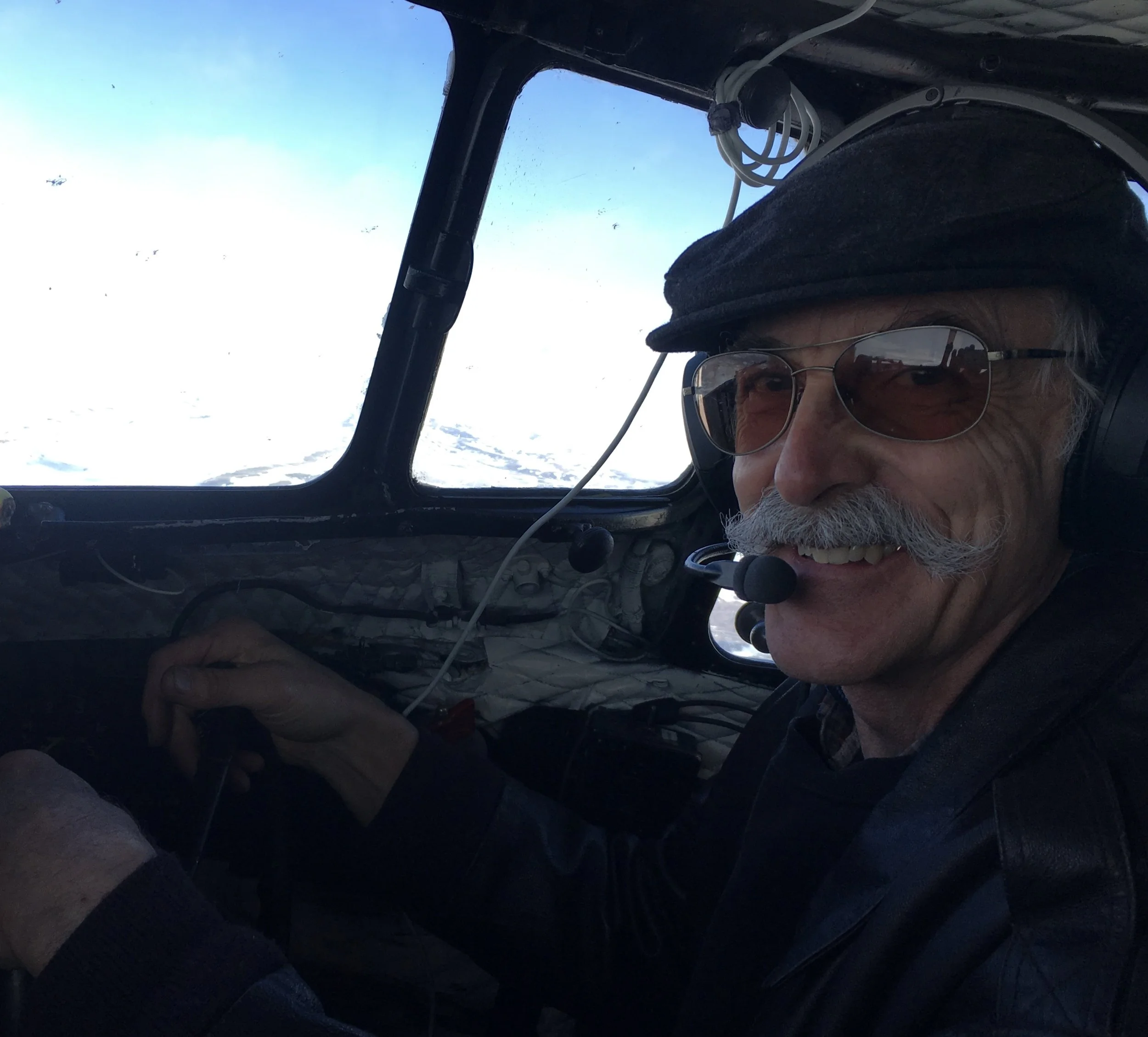 A man with a mustache and sunglasses sitting in the cockpit of an aircraft, smiling, wearing a cap and a headset with a microphone. Outside the window, snow-covered terrain is visible.