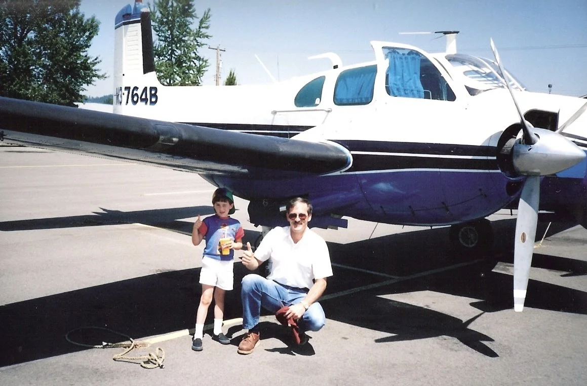 A man and a young boy posing in front of a white and blue propeller airplane at an airport, with the boy holding a snack and giving a thumbs-up.