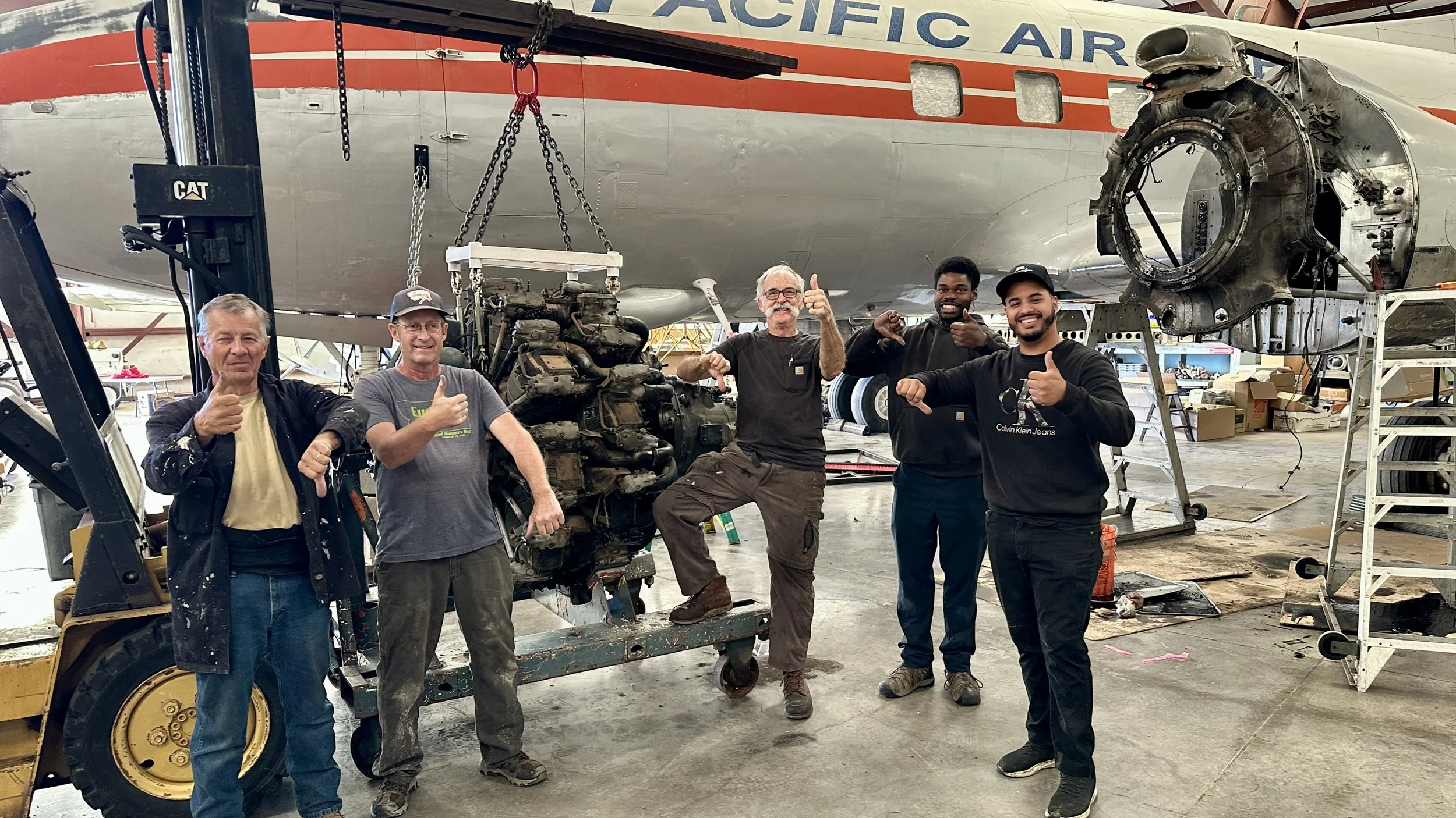 Five men standing inside an aircraft maintenance hangar, in front of a partially disassembled airplane and an engine, giving thumbs-up gestures.
