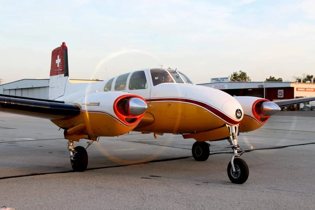 A small vintage twin-engine airplane with a white, red, and yellow paint scheme parked on an airport tarmac.