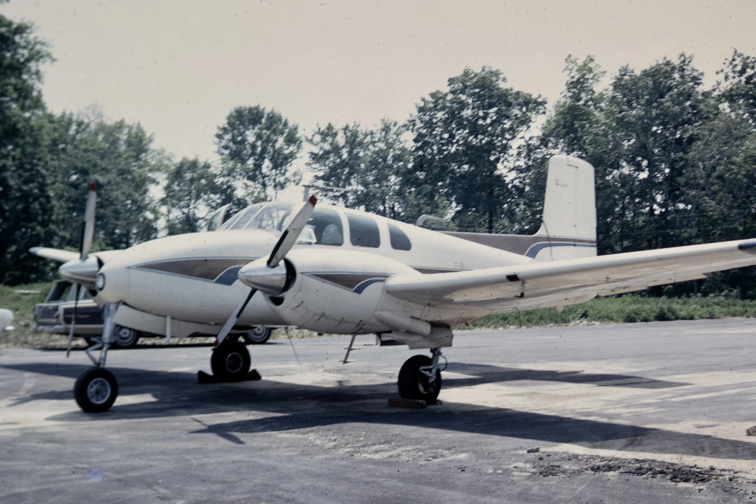 Small twin-engine military aircraft parked on tarmac with trees in the background.