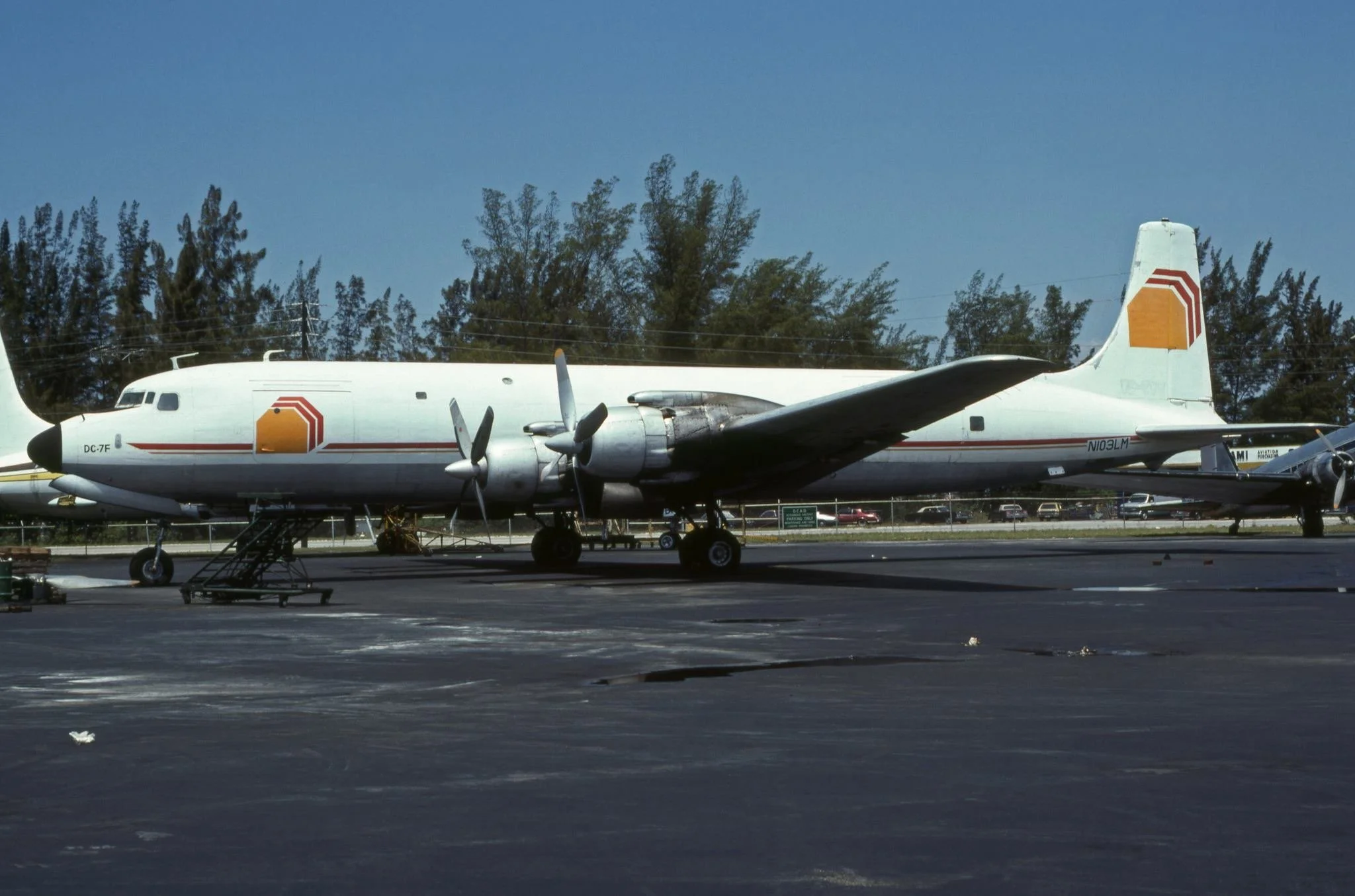 A white propeller airplane parked on tarmac with stairs leading up to it, trees and parked cars in background.