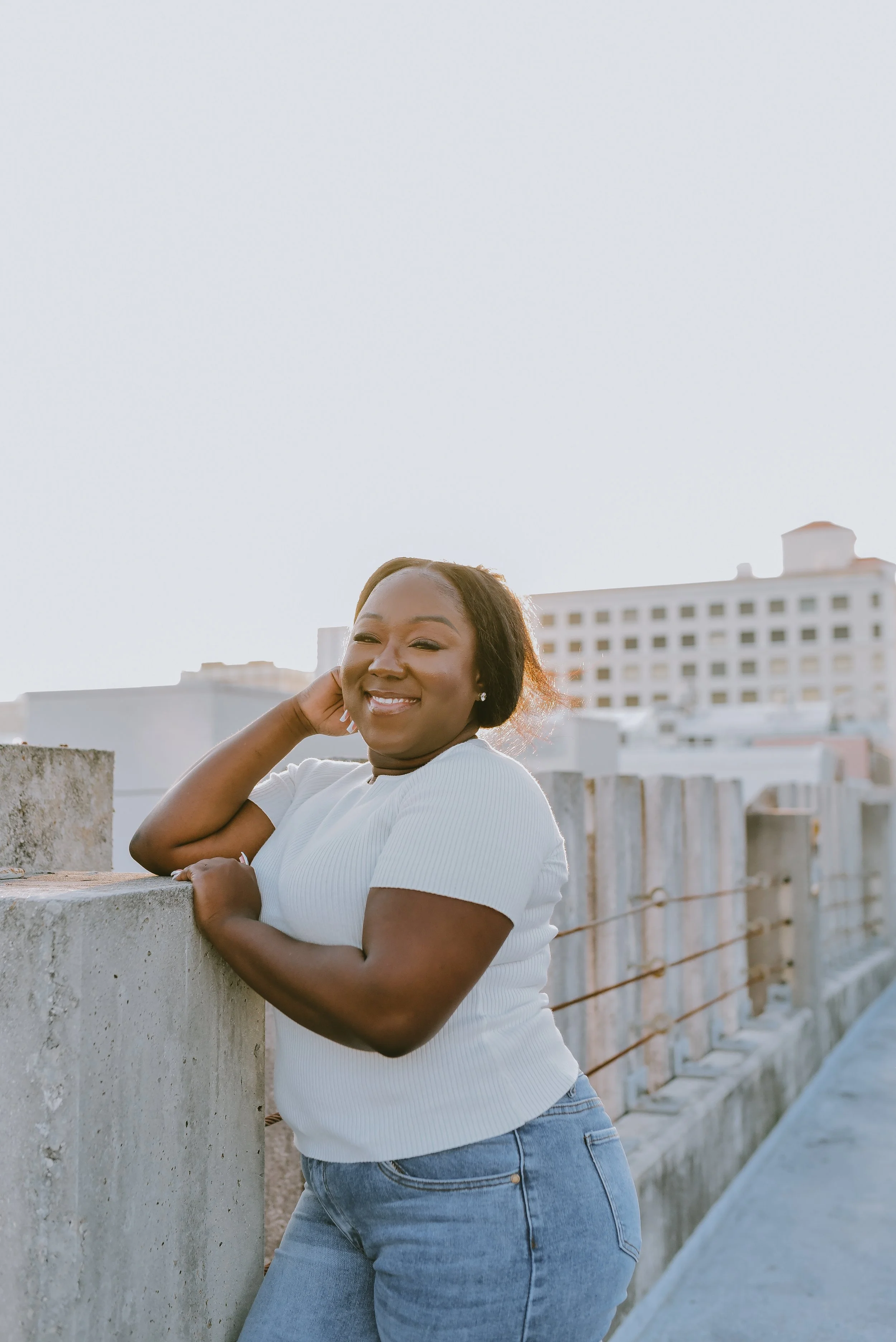 Smiling person posing outdoors near a mural and wooden fence, wearing a white shirt and jeans.