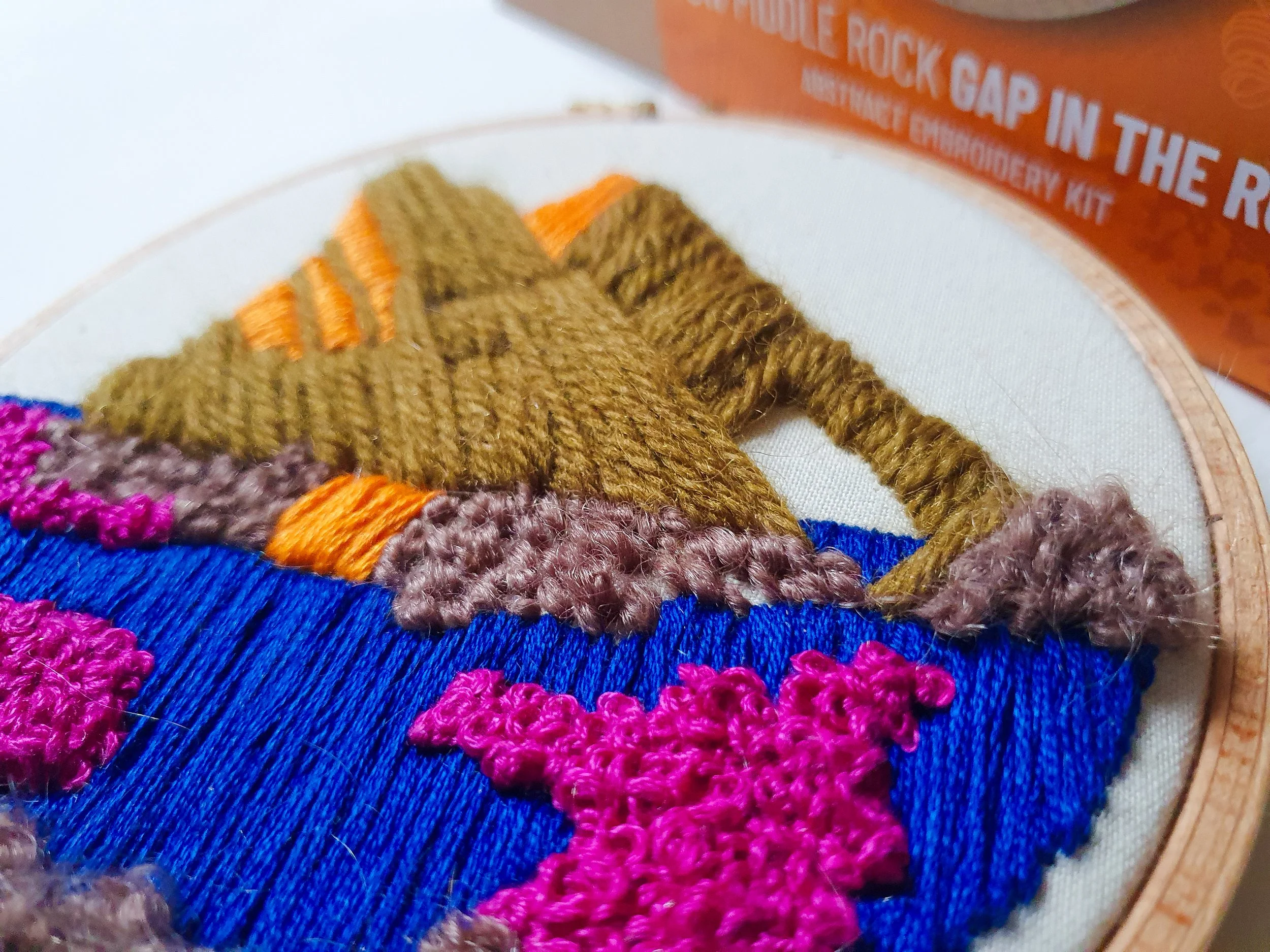 An angled image of the embroidery kit Bow Fiddle Rock 'Gap in the Rock'. Shiny satin stich in a vibrant blue with hot pink French knots and olive green tapestry yarn to complete the hoop.