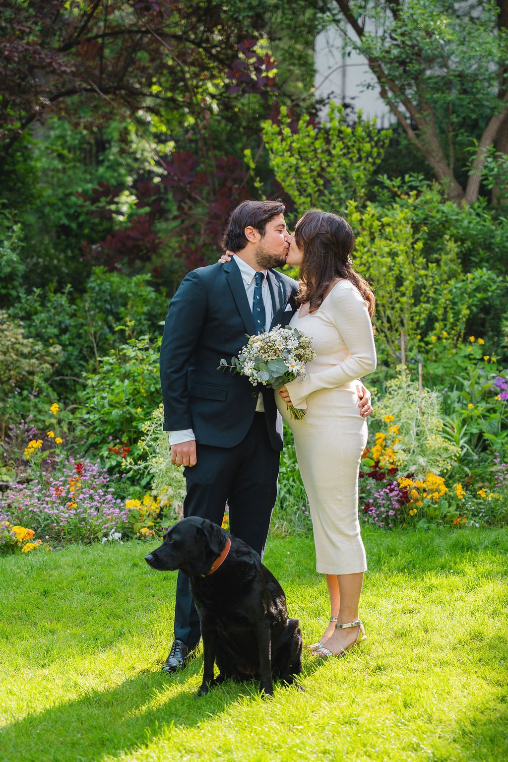 Un couple marié s'embrasse dans un jardin verdoyant, accompagné d'un chien noir assis devant eux, avec un bouquet de fleurs dans les mains de la femme.