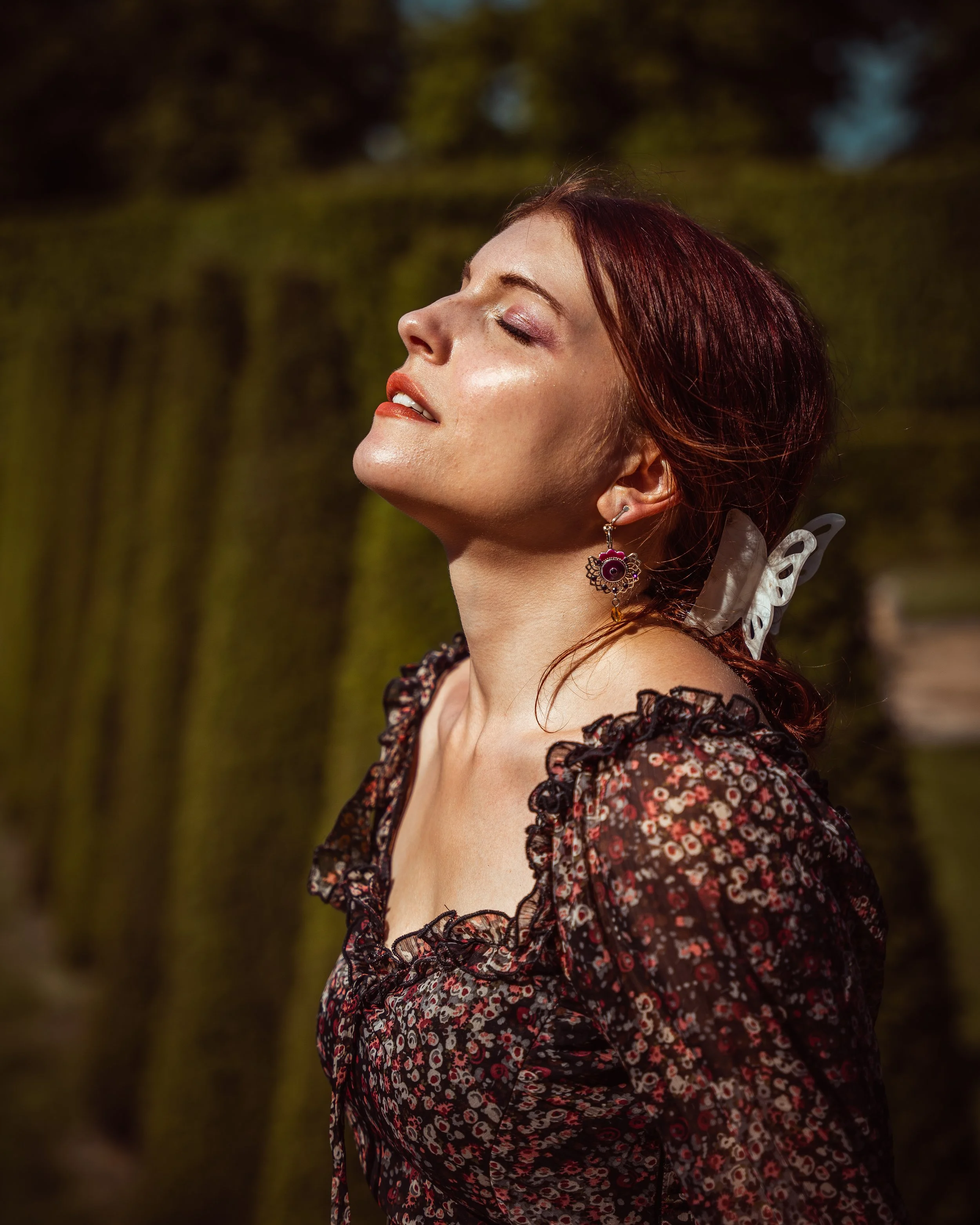 Femme avec les yeux fermés, portant un vêtement à fleurs foncées, jouant avec la lumière du soleil, dans un environnement naturel avec des arbres en arrière-plan.