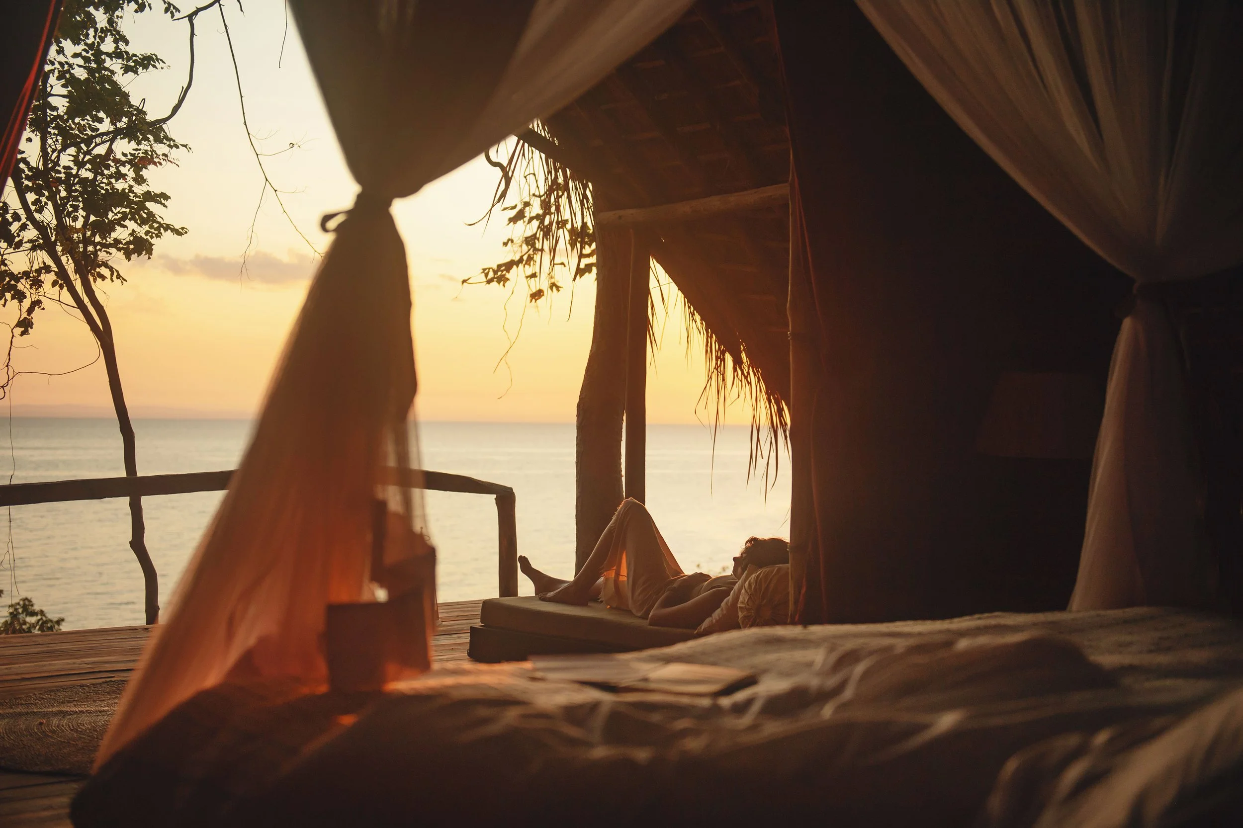 A person laying down on a bed in a rustic outdoor hut, overlooking the ocean at sunset with partly cloudy sky and trees around.