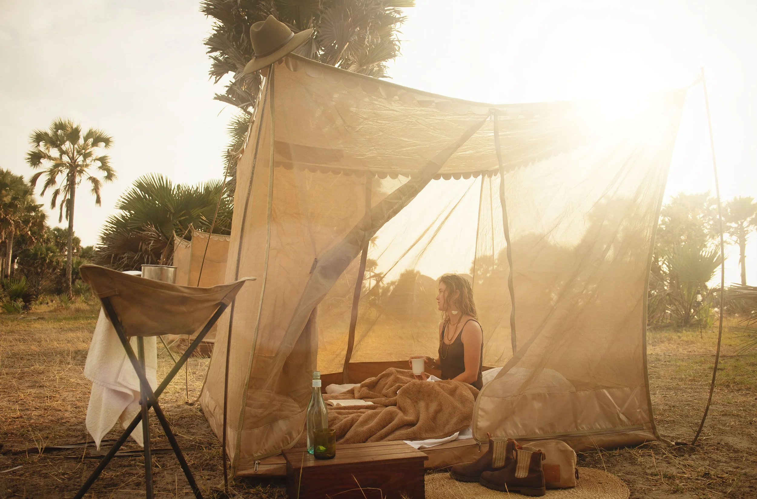 A woman sitting inside a beige outdoor tent in a desert landscape, holding a mug with mountains and palm trees in the background under a bright sun.
