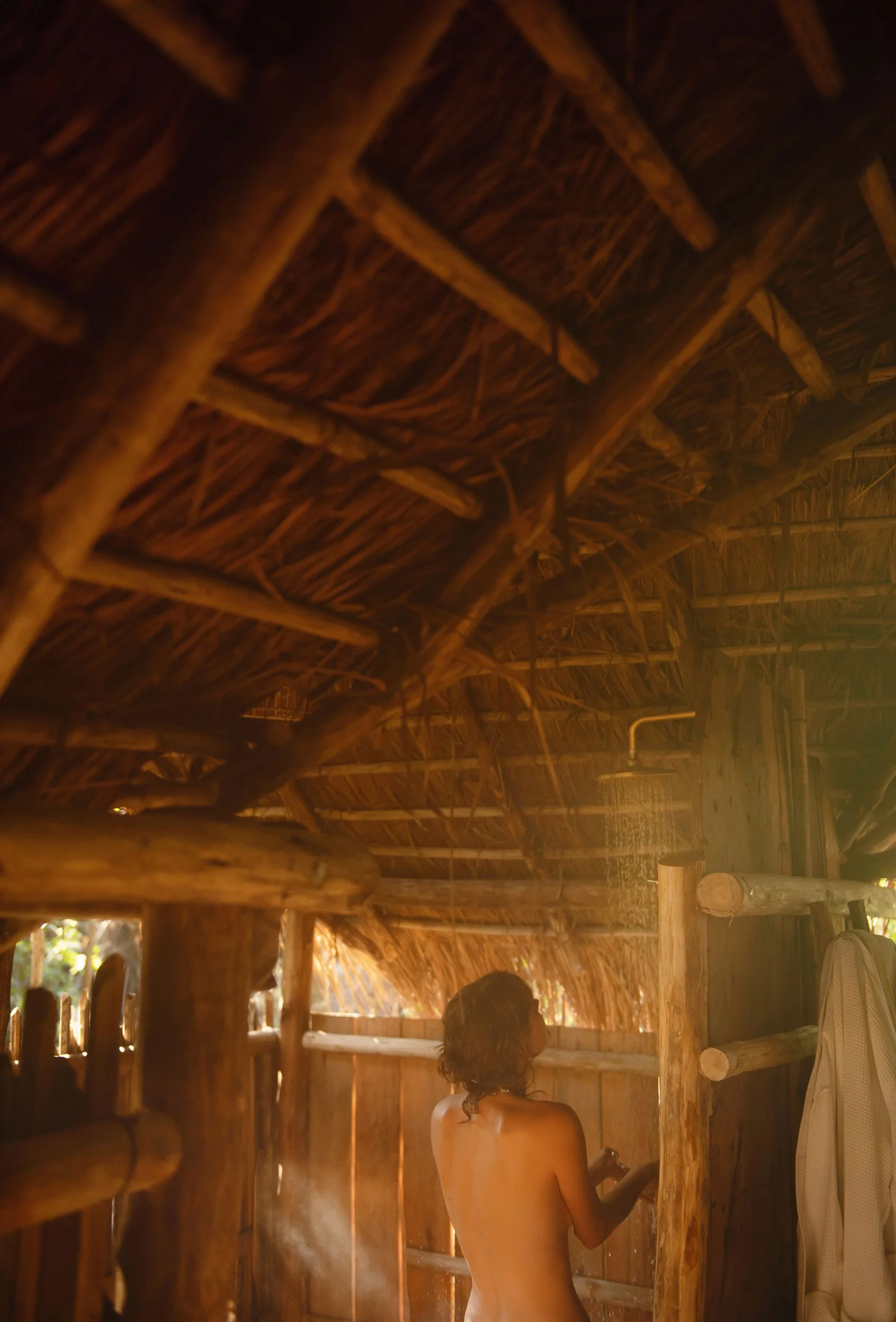 A person taking a shower in a rustic, open-air wooden hut with a thatched roof.
